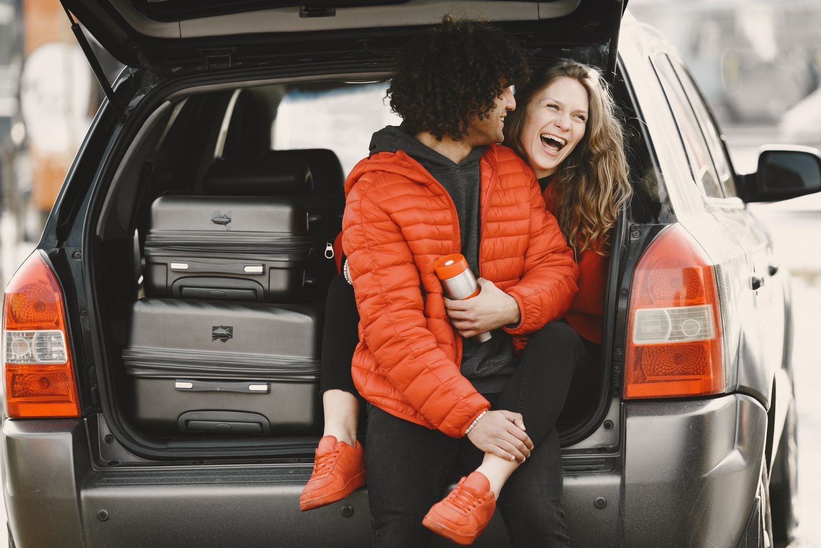 Casal sentado no porta-malas do carro cheio de malas, prontos para uma road trip